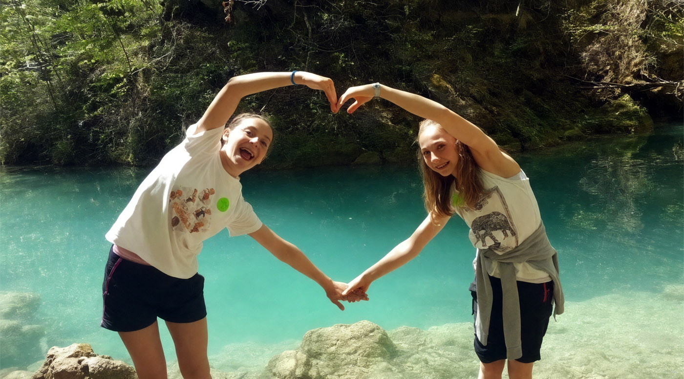 Two girls forming a heart shape with their arms by a turquoise river.